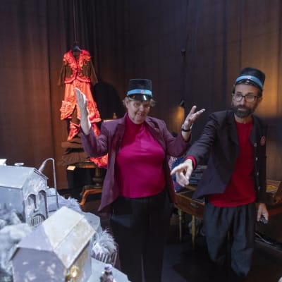 Två personer klädda som museivakter står i ett rum omgivna av scenografi och teaterrekvisita.