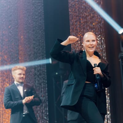 Nainen mustassa housupuvussa tuulettaa PRO-gaalan lavalla. Vaalean naisen toisessa kädessä on kuohuviinipullo. Taustalla näkyy kimaltelevia somisteita ja esiintymislavan valoja.