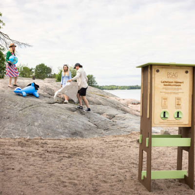 En datorbild osm visar en biljettautomat där man kan köpa biljett för Östersjöns skydd. Den finns på en badstrand med berg i bakgrunden där det sitter folk.