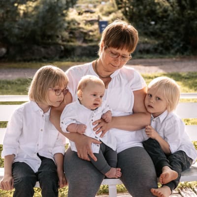 Äiti ja kolme lasta istuvat valkoisella puistonpenkillä.