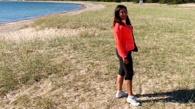 En leende kvinna med  långt mörkt hår och en orange jacka står på en lång sandstrand och ser in i kameran.