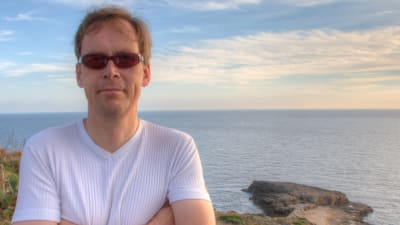 Man poserar vid strand, hav i bakgrunden. 
