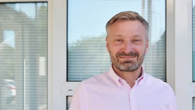 En medelålders man i rosa kragskjorta står utomhus framför ett hus.