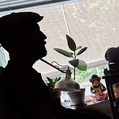 En man med keps i skägg i svart siluett sitter vid en dator med ljusa gardiner fördragna. På fönsterbrädet sitter leksaksapor med randiga byxor.