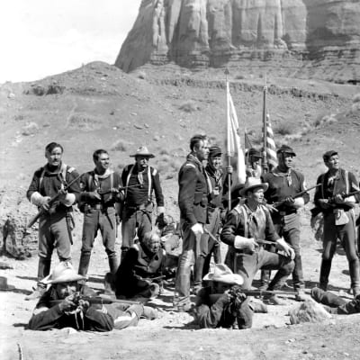 Väsyneen näköisiä USA:n ratsuväen sotilaita ryhmittyneenä lippujen ympärille autiomaassa, keskellä valkohattuinen John Wayne. Kuva elokuvasta Apassilinnake.