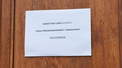 En papperslapp upphängd på en trädörr. På lappen står det: Andakt med lunch serveras i finska församlingshemmet, Lundagatan 5. Välkommen.