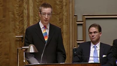 Oras Tynkkynen står i talarstolen i riksdagen och talar, han har svart kostym och regnbågsslips, snett bakom syns Jyrki Katainen.