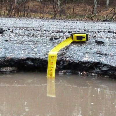 En vattenfylld grop i asfalten med ett måttband nerstucket. Måttbandet visar på lite under sju centiometer vatten.