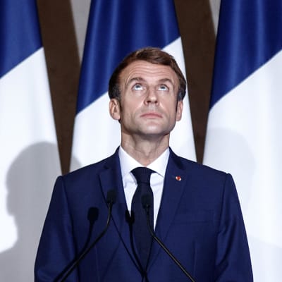 Emmanuel Macron står vid ett talarpodium. Han tittar uppåt. Han är klädd i en mörkblå kostym och svart slips. I bakgrunden Frankrikes flagga och EU:s flagga.