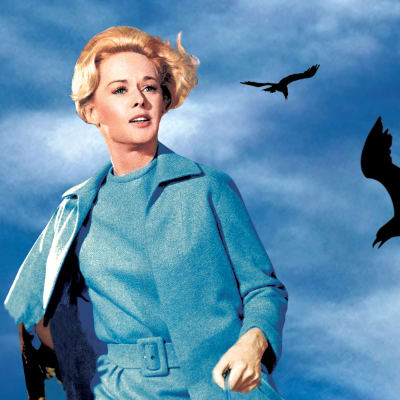 Hätääntyneen näköinen nainen (näyttelijä Tippi Hedren) pakenee tukka hulmuten, taustalla mustia lintuja taivaalla. Hitchcockin elokuvan Linnut mainoskuva.