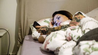 En ung kvinna ligger i sängen och tittar på sin mobiltelefon.