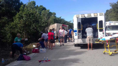 Sjukvårdspersonal hjälper skadade och chockade turister vid en vägren efter en bussolycka. Olycksbussen i bakgrunden har vält och ligger på sida vid sidan om vägen.