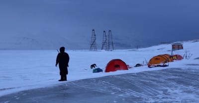 En person står bredvid några tält. Det ser ut som om hen håller vakt. Runt omkring syns snö och berg. 