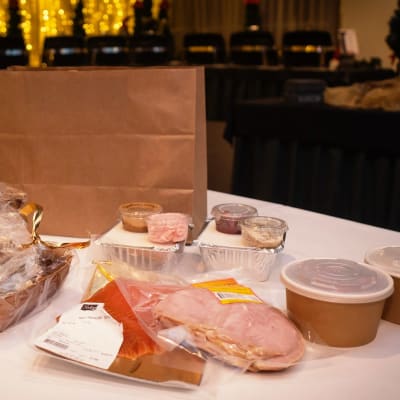 Paperikassi, jonka edessä on purkkeihin pakattua jouluruokaa, vakuumipakattua kinkkua- ja kalaa sekä kasa leivonnaisia.