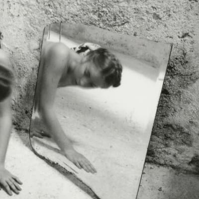 Mustavalkoinen valokuva, jossa taiteilija konttaa alastomana peilin eteen.