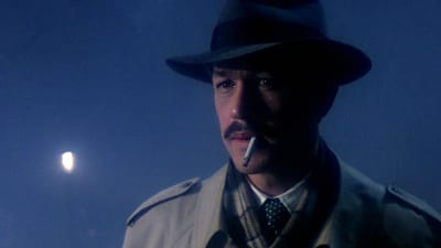 Hattuun ja trenssitakkiin pukeutunut tupakoiva mies (näyttelijä Frederic Forrest) puolilähikuvassa, taustalla sinistä sumua ja katulyhty. Kuva elokuvasta Hammett - tehtävä Chinatownissa.