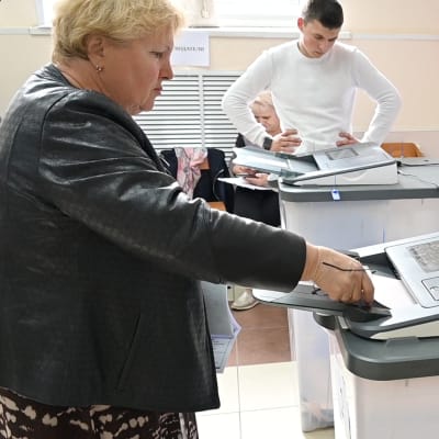 Vaaleahiuksinen nainen mustassa takissa syöttää äänestyslippua sähköiselle äänestyslaitteelle. Taustalla nuori mies valkoisessa paidassa tarkastelee vastaavaa konetta kädet lanteillaan. Seinällä on vaalijuliste.