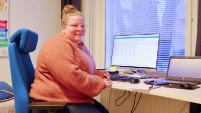 En kvinna i orange tröja sitter framför ett skrivbord med två datorskärmar. Hon ler och tittar in i kameran. Bakom henne syns en bäddad säng.