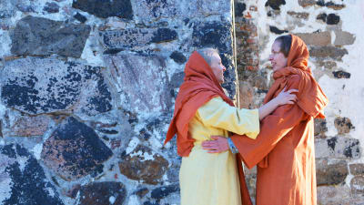 Två kvinnor i långa klänningar och huvuddukar håller om varandra och ser glada ut. Det är två skådespelare som spelar Johanna från Galileen och Maria från Magdalena. Bakom dem synns en stenvägg i solljus.