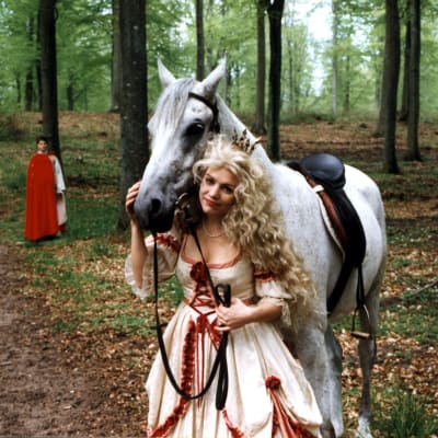 En blond flicka står vid sidan och håller en vit häst.