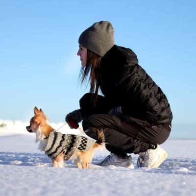 Jonna Löfgren sitter på huk i ett vintrigt landskap. Hon har svarta kläder och grå mössa. Bredvid henne står hennes lilla hund Sisu som har en Malaxtröja för hundar på sig.