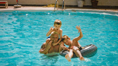 Paul med sina två små barn i en bassäng med turkost vatten. Ena barnet sitter i en simring och det andra på Pauls axlar.