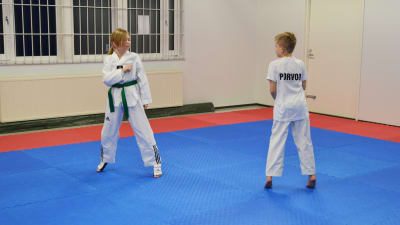 Två barn i vita dräkter står mitt emot varandra. Det ser ut som om de ska påbörja en taekwondomatch. De står i en idrottssall med rödblå gummimatta på golvet.