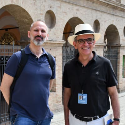 Två män tittar leende in i kameran invid en utomhusopera i  Macerata, Italien.