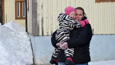 Porträttfoto av kvinna med barn i famnen. Kvinnan har svart jacka, mörkt hår och blå jeans. Barnet är 2,5 år gammalt och har en zebrarandig overall samt en rosa mössa. I bakgrunden syns ett hus och snö.