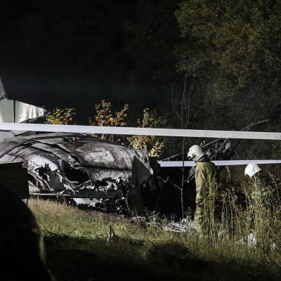 Två personer i hjälm och räddningsoverall står bredvid ett totalförstört flygplan.