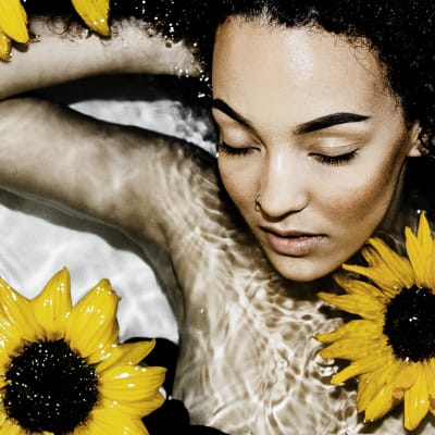Nainen makaa vesialtaassa silmät kiinni pidellen toista kättään päänsä takana. Hänellä on mustat afrokiharat. Vedessä kelluu auringonkukkia.