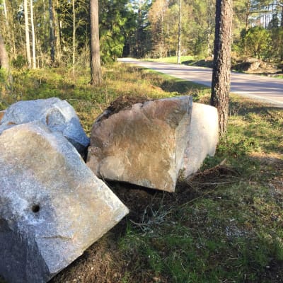 Tre jättestora stenblock ligger vid vägkanten vid en asfalterad väg i skogen.