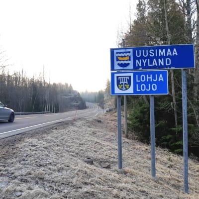 En skylt där det står Nyland och en annan där det står Lojo vid en väg. På vägen kör en bil.