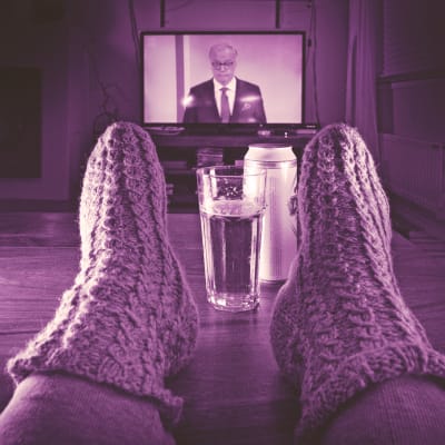Televisio auki jossa näkyy tv-uutiset, katsoja kotona sohvalla jalat pöydällä ja pöydällä olutlasi.