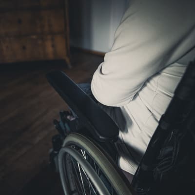 Valkoiseen huppariin pukeutunut ihminen istuu pyörätuolissa, lähikuva ylhäältäpäin henkilön olkapäästä ja pyörätuolin käsinojasta.