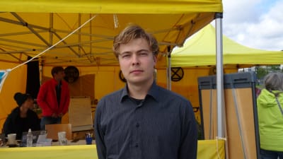 Amos Wallgren står utanför ett gult tält på medborgarplatsen i Helsingfors. Han tittar rakt in i kameran och är klädd en mörkblå kragskjorta.