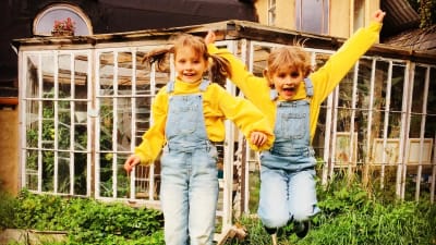 Två likadant klädda flickor hoppar upp i luften ute i en somrig trädgård