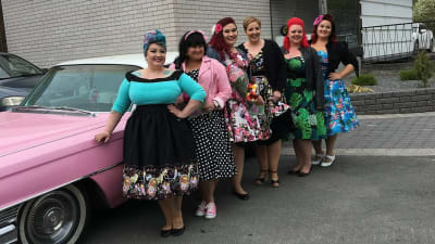 Sex kvinnor klädda i vintage-stil på parkeringsplats tillsammans med chockrosa bil från 1964.