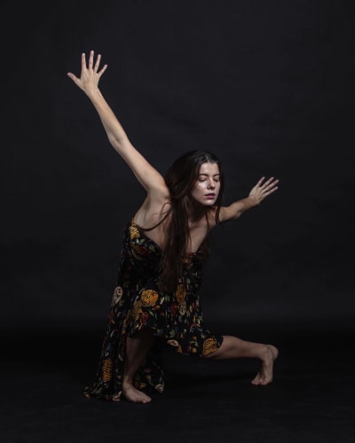 Balettitanssija Suvi Honkanen kirjavassa painokuosisessa olkaimettomassa mekossa poseeraa vasten mustaa taustaa vasten.