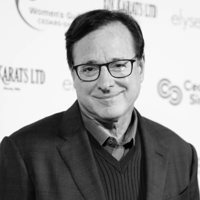 Svartvit bild av Bob Saget som poserar på röda mattan. Han tittar in i kameran och ler.