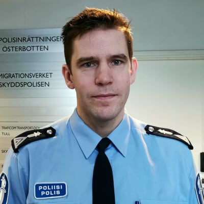 En man i polisuniform står intill en vit vägg. På väggen finns ett plakat med texten polisinrättningen i Österbotten.