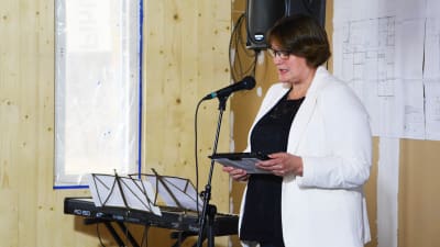 En medelålders kvinna i vit kavaj står framför en mikrofon och läser från någonting.