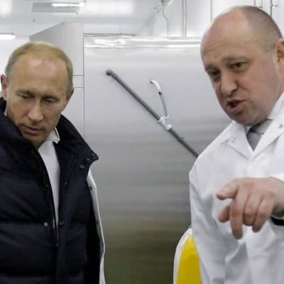 Vladimir Putin och Jevgenij Prigozjin i ett storkök. Putin har ett vitt skyddsplagg över en mörk jacka och Prigozjin som pekar på något utanför bild är helt klädd i vitt.