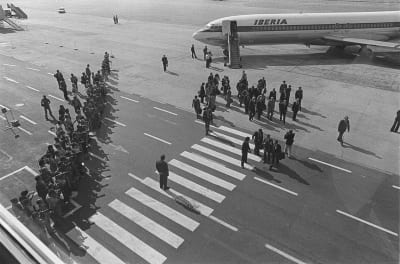 Ulkomaisen vieraan vastaanotto: presidentti Urho Kekkonen kävelee vieraineen kohti lentoasemaa, ympärillä parveilee valokuvaajia, valokuvaajien rivi köyden takana.