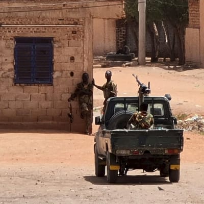 Soldater i sudaneiska armén tar en paus vid ett gathörn. En soldat lutar sig mot väggen medan en annan står bredvid. En tredje soldat sitter vid ett maskingevär på flaket till ett militärfordon.