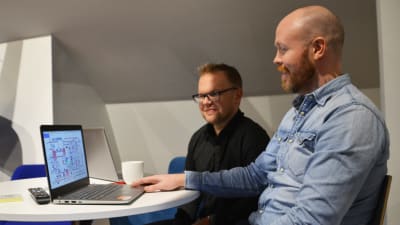 Två män sitter vid ett bord och tittar på en bärbar dator.