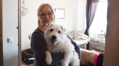En medelålders kvinna står inne i en lägenhet. Hon håller en vit hund i famnen och båda tittar in i kameran.
