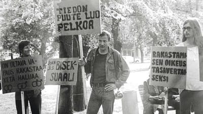Mustavalkoinen kuva, jossa nuoria mielenosoittajia iskulausekylttien kanssa ja keskellä nuori Veli Hyvärinen.