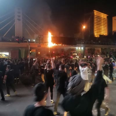 Någon håller upp en fackla under en demonstration i Teheran. En stor folksamling har samlats på en öppen plats i kvällsbelysning med en bro och höghus i bakgrunden.