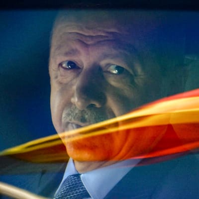 Pääministeri Recep Tayyip Erdogan katsoo suoraan kameraan, välissä on lasi jossa näkyy värillisiä heijastuksia.